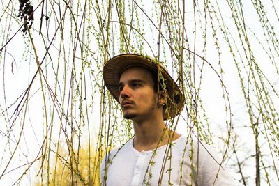 Thoughtful young man wearing hat while standing amidst long hanging branches