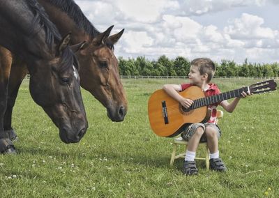 Full length of boy holding guitar by horses on grassy field