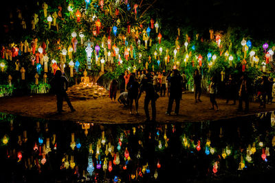 People with multi colored lanterns on tree at lakeshore during night