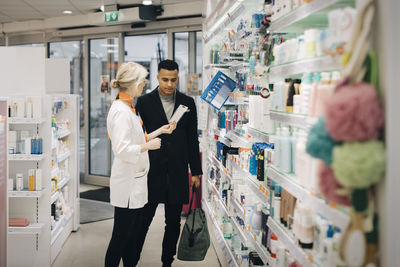 Side view of mature female pharmacist assisting male customer standing by rack at medical store