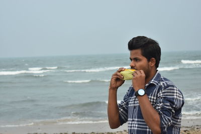 Young man eating food at beach against sky