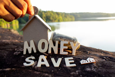Cropped hand of man holding coin by model home with money save text on rock by lake