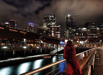 Rear view of woman standing on bridge by illuminated buildings in city at night