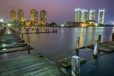 Pier on river by illuminated buildings against sky at night