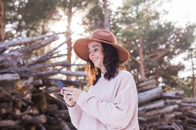 Smiling woman using smart phone while standing against woodpile in forest