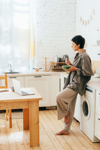 Young attractive woman eats salad while standing in the kitchen and watching video on laptop.