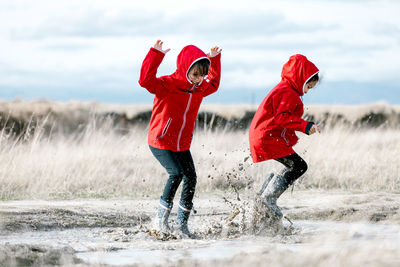 Carefree kids in raincoats and rubber boots splashing water in puddle while having fun and laughing on sunny day in countryside