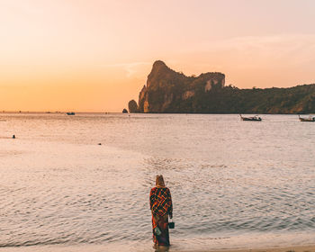 Rear view of woman standing at beach against sky during sunset