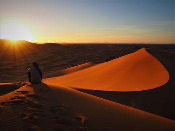 Rear view of man sitting at desert against sky during sunset