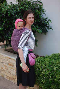 Portrait of young woman carrying child in wrap on her back standing against plants