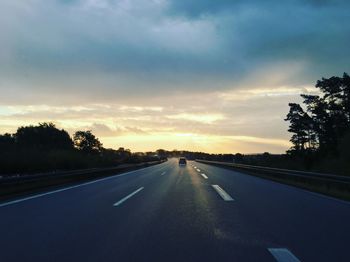 Empty road along trees and against cloudy sky