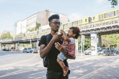 Young man carrying daughter holding mobile phone while standing on street against railway bridge