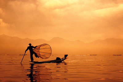 Silhouette man fishing in sea while standing on boat at sunset