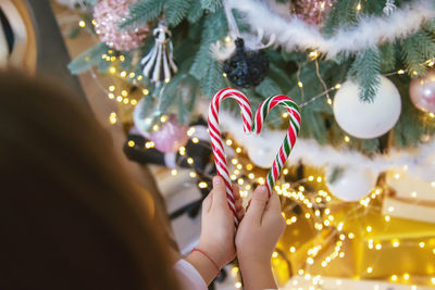 Cropped hand of girl holding candy cane against christmas tree at home