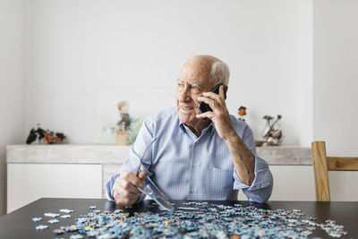 Happy senior man using smartphone while doing a puzzle at home