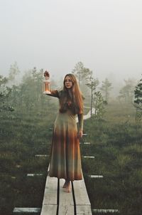 Full length of woman holding lantern while standing on boardwalk during foggy weather