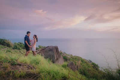 Rear view of man standing on rock by sea against sky during sunset