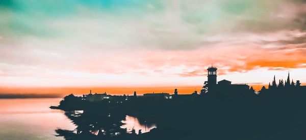 Silhouette buildings by sea against sky during sunset