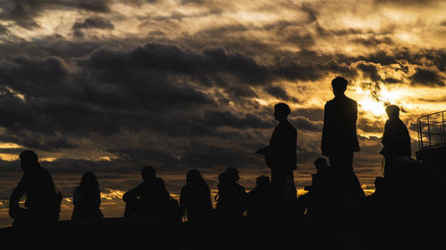 Back view of unrecognizable silhouette of a group of tourists sitting and standing on roof of building looking at view of city sunset