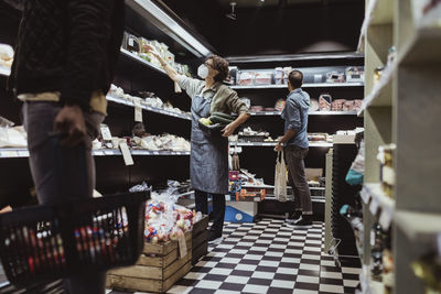 Male customers collecting package product while female owner standing by arranging food on rack at deli store during pan