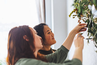 Female friends decorating christmas wreath on wall at home 