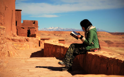 Side view of young woman reading book while sitting on retaining wall against sky