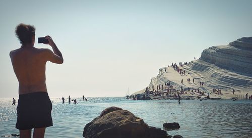 Rear view of shirtless man standing on rock by sea against clear sky