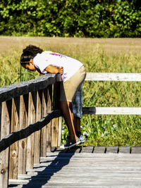 Side view of girl leaning on wooden railing over field
