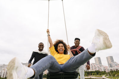 Happy men pushing friend sitting on swing in park