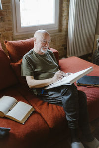 Senior man doing painting while sitting on sofa by book at home