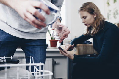 Midsection of man holding cup and glass in kitchen with woman using mobile phone at home