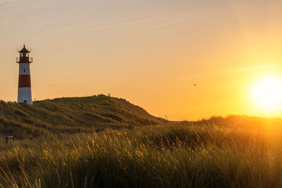Lighthouse on field by buildings against sky during sunset