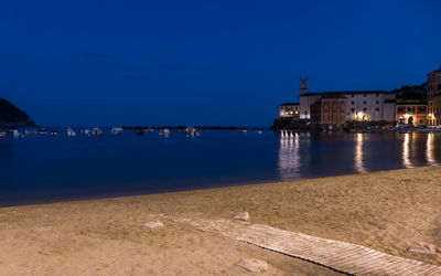 Illuminated buildings by sea against blue sky at night