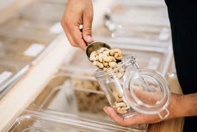 Side view of anonymous female pouring cashews inside glass jar in local store