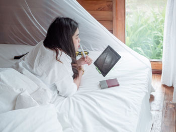 Young asian woman freelance working on notebook and tablet remotely while rest in bedroom