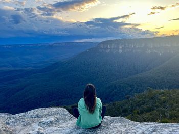 Rear view of woman sitting on mountain against sky during sunset
