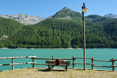 Rear view of woman sitting on bench by lake against mountains