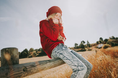 Woman sitting on railing in field against sky
