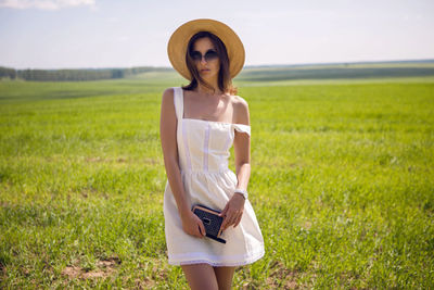 Young woman with long black hair stands in white dress and clothes straw hat is on the green field 