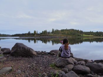 Rear view of woman sitting on rock by lake against sky