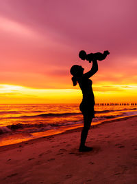 Young adult woman mother holding baby in air on beach with sunset background silhouette vertical