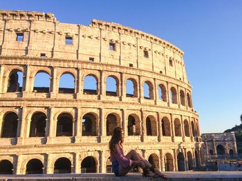 Side view of woman sitting on retaining wall against coliseum