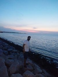 Man standing on rock by sea against sky during sunset