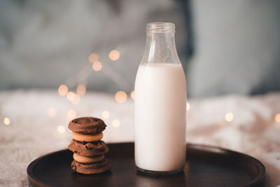 Glass bottle with fresh milk and stack of chocolate cookies on wooden tray in bed closeup. 