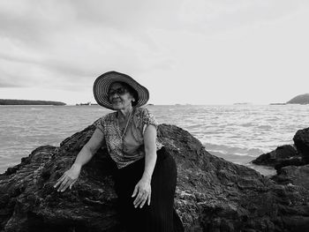 Senior woman wearing hat sitting on rock by sea against sky