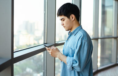 Side view of young man standing against window