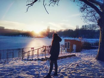 Rear view of woman standing on snow covered landscape by frozen lake against sky during sunset