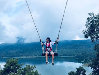 Man on swing with lake in background against cloudy sky