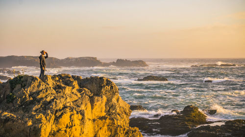 Side view of man standing on cliff by sea against sky during sunrise