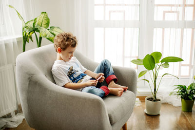 A cute boy is sitting in an armchair and playing a game on his phone or watching educational videos.